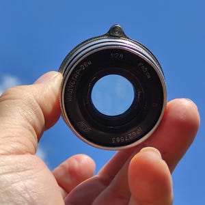 May include: A vintage camera lens with black and silver metal casing. The lens has the text "ИНДУСТАР-26м" and "F=5 cm" printed on it. The lens is held against a bright blue sky.