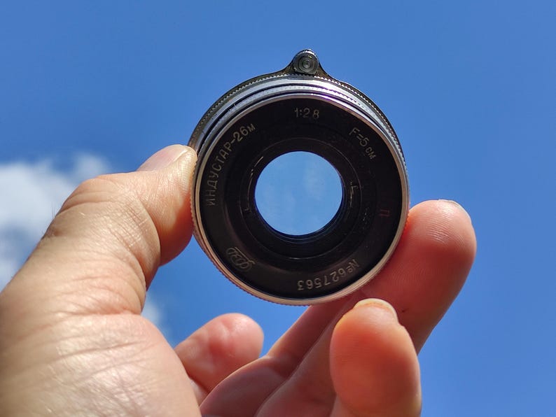 May include: A vintage camera lens with black and silver metal casing. The lens has the text "ИНДУСТАР-26м" and "F=5 cm" printed on it. The lens is held against a bright blue sky.
