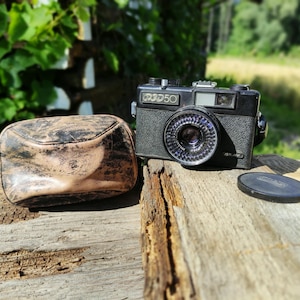 Peut inclure: Un appareil photo noir FED 50 avec un bouchon d'objectif et un étui en cuir marron. L'appareil photo est posé sur une surface en bois. L'objectif présente un motif circulaire. L'étui est marron avec un aspect vieilli.