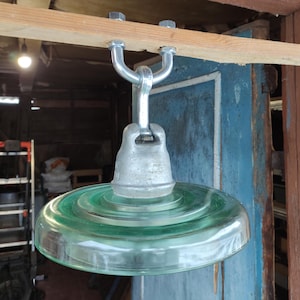 May include: A green glass insulator with a metal mounting bracket. The insulator is hanging from a wooden beam.