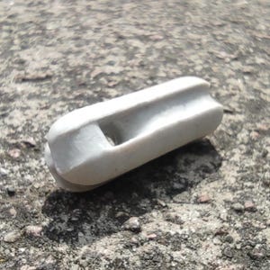 May include: A white ceramic insulator with a rectangular body and a central hole. The insulator has rounded edges and is designed for electrical applications. It is resting on a rough, grey surface.