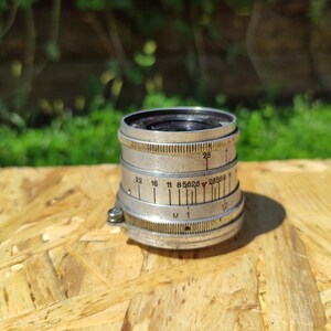 May include: A vintage silver camera lens with black details and numerical markings. The lens is cylindrical with ridged edges and a small screw. It sits on a wooden surface with a blurred green background.