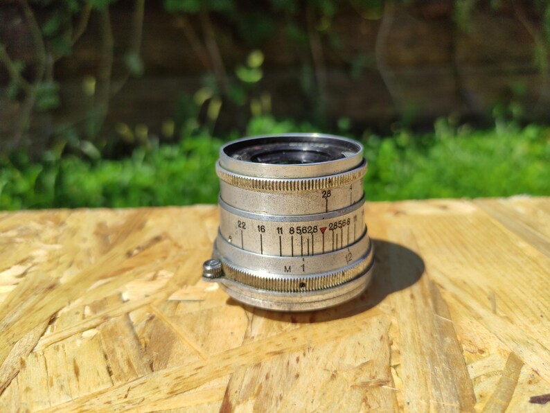 May include: A vintage silver camera lens with black details and numerical markings. The lens is cylindrical with ridged edges and a small screw. It sits on a wooden surface with a blurred green background.