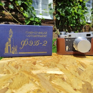 Puede incluir: Una cámara vintage en una funda de cuero marrón está junto a una caja azul con letras doradas. La cámara es plateada y negra con una lente grande. La caja tiene texto ruso y un gráfico de un monumento.