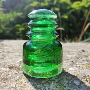 Puede incluir: Un aislador de vidrio verde esmeralda vintage con una parte superior redondeada y un diseño acanalado. La pieza antigua se exhibe al aire libre, con un fondo verde borroso. El vidrio tiene un acabado brillante y es parcialmente transparente.