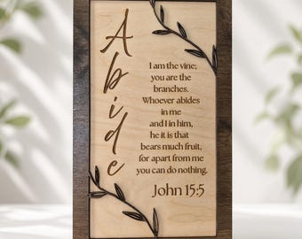 May include: A rectangular wooden wall sign featuring a dark brown frame and a light wood centre panel. The word Abide is written vertically in large script, accompanied by engraved text from John 15:5 and decorative dark vine accents.