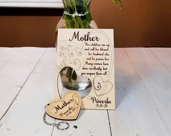 May include: A wooden heart-shaped keyring with the text "Mother Proverbs 31:28-29" on it. The keyring is hanging from a silver chain. The keyring is in front of a wooden sign with the text "Mother Her children rise up and call her blessed: her husband also. and he praises her: Many women have done excellently, but you surpass them all. Proverbs 31:28-29". The sign is in front of a vase with a bouquet of pink, yellow, and green flowers.