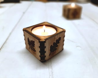 May include: A square wooden candle holder with leaf designs. A white candle is burning inside the holder.