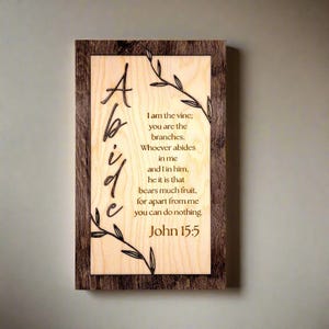 May include: A wooden sign with a dark brown frame and a light wood background. The sign features a laser-cut design of a vine with leaves and the text "Abide" in a decorative font. The text "I am the vine; you are the branches. Whoever abides in me and I in him, he it is that bears much fruit, for apart from me you can do nothing. John 15:5" is also laser-cut onto the sign.