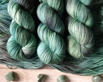 May include: Three skeins of hand-dyed yarn in shades of teal, green, and gray, with a speckled appearance. The yarn is arranged with several polished green gemstones on a light wood surface. The yarn is suitable for knitting or crochet projects.
