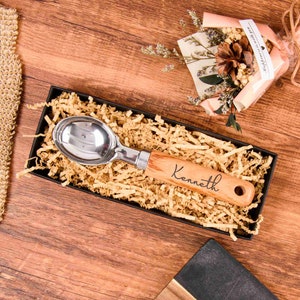 May include: A personalised wooden ice cream scoop with a silver metal scoop and the name "Kenneth" engraved on the handle. The scoop is in a box with brown paper shreds.