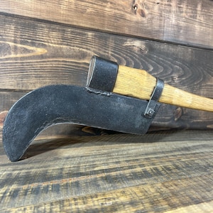 May include: A large, curved blade tool with a wooden handle. The blade is black and crescent-shaped, attached to a wooden handle with metal bands. The handle is light brown, and the tool rests on a wooden surface.