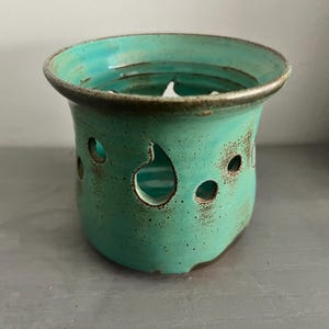 May include: A teal ceramic candle holder with cut-out designs. The holder has a distressed finish and a brown rim.