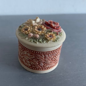 May include: A small, round, white trinket box with a floral design. The lid is decorated with a cluster of yellow, pink, and red flowers. The box has a red and white floral pattern around the bottom.