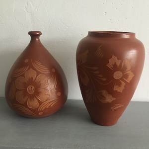 May include: Two terracotta vases with floral designs carved into the surface. The vases are brown and have a rustic look.