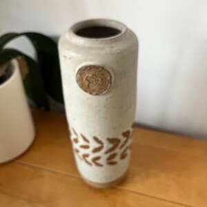 May include: A cylindrical ceramic vase with an off-white speckled glaze. The vase features a circular medallion detail near the top rim and a repeating brown leaf-like pattern encircling the lower section of the body, finished with a smooth rim.