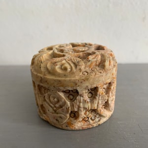 May include: A round, intricately carved stone trinket box with a floral design. The box is made of light brown and beige stone with hints of orange and red.