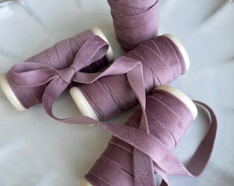Puede incluir: Varios carretes de cinta de terciopelo rosa empolvado están colocados sobre un plato blanco con borde festoneado. La cinta está enrollada alrededor de carretes de madera, con algunos carretes desenvueltos y atados en lazos. El color es un púrpura apagado.