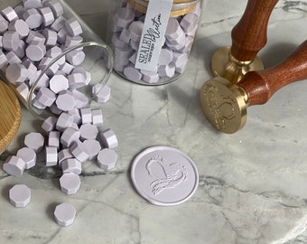 May include: A collection of lavender wax seal beads in a glass jar with a bamboo lid, alongside a fallen jar of beads, a stamped seal, and two wooden handled wax seal stamps. The jar has a tag that says "SEALED COLLECTION".