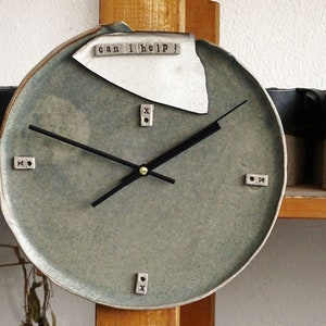 May include: A round, green ceramic clock with black hands and white ceramic accents. The clock face has the words "can I help?" written on it.