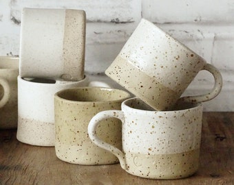 Peut inclure: Cinq mugs en céramique avec un glaçage moucheté dans des tons de blanc et beige. Les mugs sont empilés et disposés sur une surface en bois.