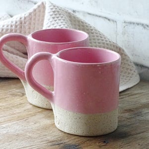 Op de afbeelding: Twee keramische mokken met een tweekleurig ontwerp, voorzien van een glanzend roze bovenzijde en een gespikkelde, ongeglazuurde beige basis. Elke mok heeft een gebogen handvat en een kleine cirkelvormige stempel nabij de onderrand.