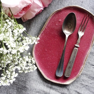 Puede incluir: Un plato de cerámica rosa con una superficie rugosa y texturizada. El plato tiene una forma ligeramente ovalada y presenta una cuchara y un tenedor de plata.
