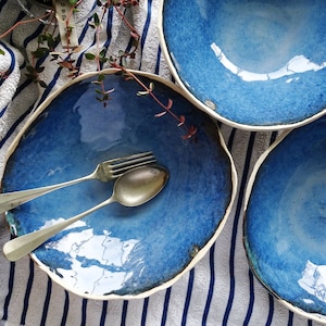Może przedstawiać: Trzy ręcznie wykonane miski ceramiczne o nieregularnym, organicznym kształcie i błyszczącej, głęboko niebieskiej glazurze. Miski mają biały, nieszkliwiony brzeg i towarzyszy im srebrny metalowy widelec oraz łyżka umieszczone wewnątrz.