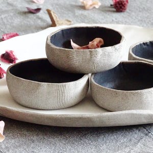 Può includere: Tre piccole ciotole rotonde in ceramica con un esterno grigio chiaro testurizzato e un interno nero opaco liscio. Le ciotole presentano un sottile motivo lineare sulla superficie esterna e sono disposte insieme su un piatto in ceramica irregolare.