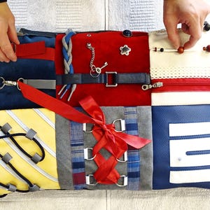Op de afbeelding: Een rechthoekig sensorisch activiteitskussen met een patchworkontwerp van verschillende stoffen en texturen. Het bevat een rode riem, een blauw gevlochten koord, een metalen sleutel aan een ketting, een rode lintvetersluiting, een rits en houten kralen aan een touwtje.