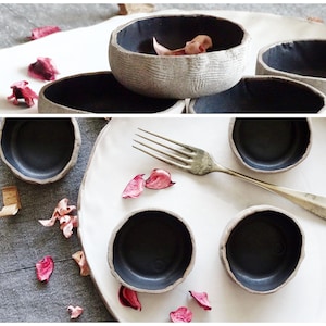 Pode incluir: Conjunto de tigelas de cerâmica artesanais, com exterior cinza texturizado e interior escuro. Dispostas sobre uma superfície branca, com um garfo e pétalas de flores secas. As tigelas têm um aspeto rústico e artesanal.