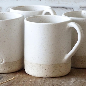 May include: Four white ceramic mugs with speckled glaze and a brown speckled band around the bottom. The mugs have a rounded shape and a handle on each side.