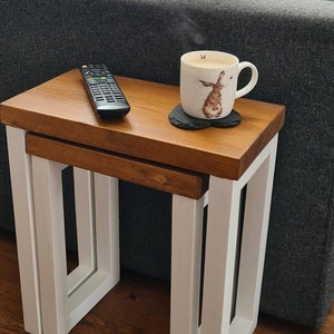 May include: A set of two nesting tables with a light brown wooden top and white painted legs. A remote control and a mug with a rabbit design are on the top table. The tables are on a hardwood floor.
