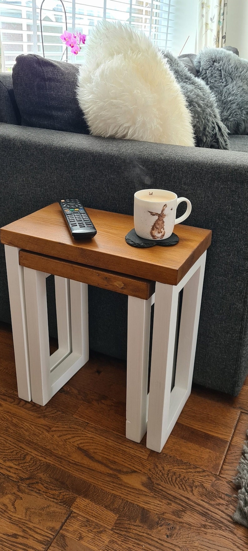 May include: A set of two nesting tables with a light brown wooden top and white painted legs. A remote control and a mug with a rabbit design are on the top table. The tables are on a hardwood floor.