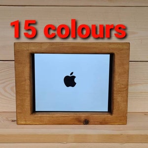 May include: A wooden frame with a white background and a black Apple logo in the centre. The frame is on a wooden surface. The text "15 colours" is above the frame.