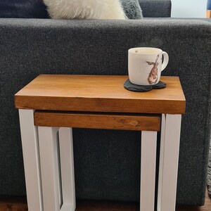 May include: A set of two nesting tables with a light brown wooden top and white legs. A white mug with a rabbit design sits on a black coaster on the larger table. The tables are in front of a gray sofa.