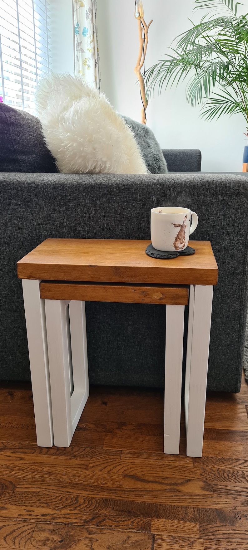 May include: A set of two nesting tables with a light brown wooden top and white legs. A white mug with a rabbit design sits on a black coaster on the larger table. The tables are in front of a gray sofa.