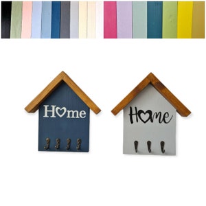 May include: Two wooden key holders shaped like houses with the word "Home" and a heart on the front. The house on the left is painted blue with white lettering and the house on the right is painted gray with black lettering. Both houses have four hooks on the bottom.