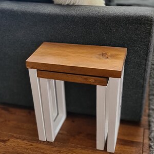 May include: A set of two nesting tables. The tabletop is a warm brown, and the legs are white. The tables are made of wood and have a modern design. The larger table is 18 inches wide, 14 inches deep, and 18 inches tall.