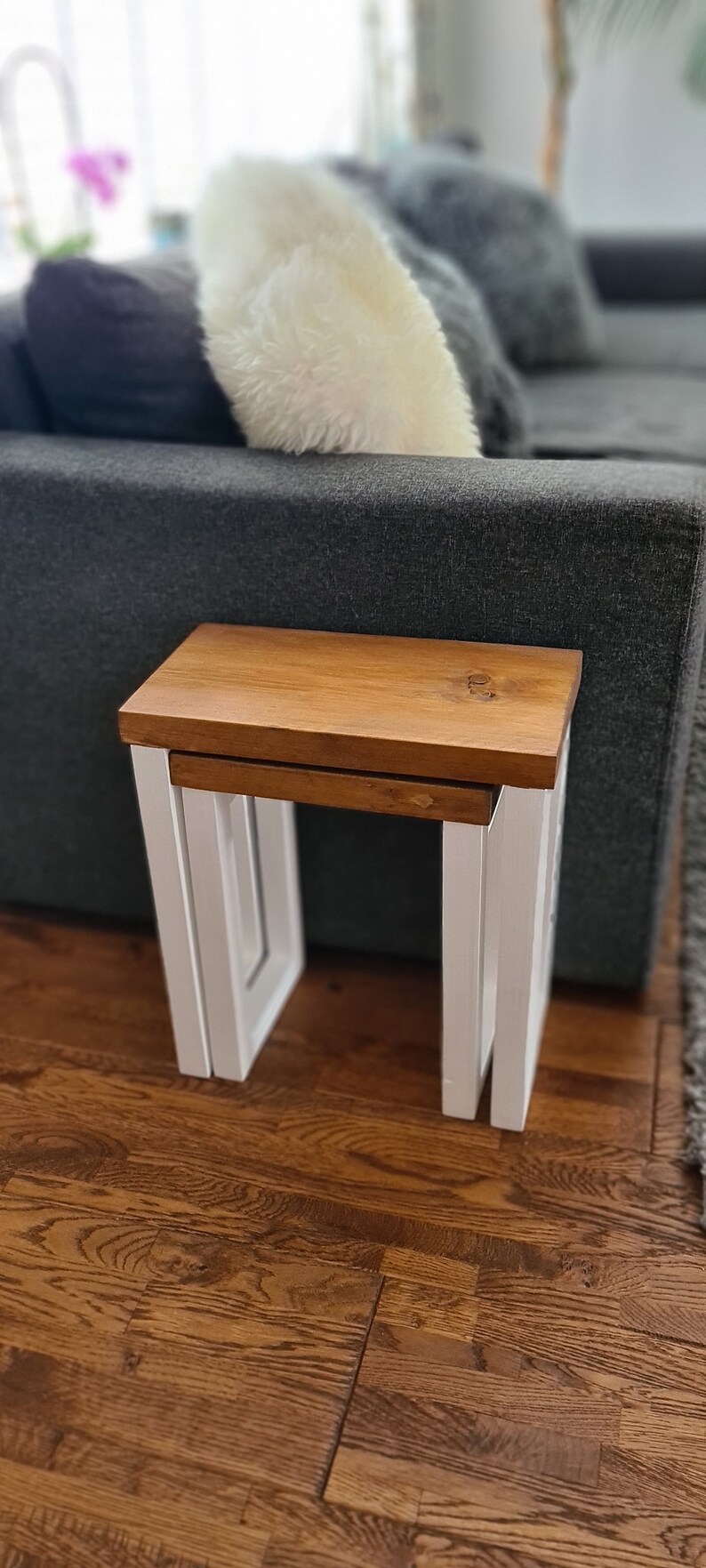 May include: A set of two nesting tables. The tabletop is a warm brown, and the legs are white. The tables are made of wood and have a modern design. The larger table is 18 inches wide, 14 inches deep, and 18 inches tall.