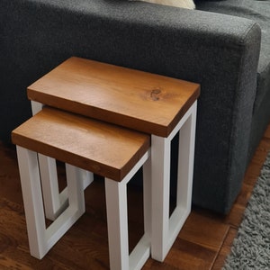 May include: Two wooden nesting tables. The tabletops are a warm brown, and the legs are white. The tables are rectangular and have a simple, modern design. The larger table is approximately 18 inches wide and 18 inches deep.