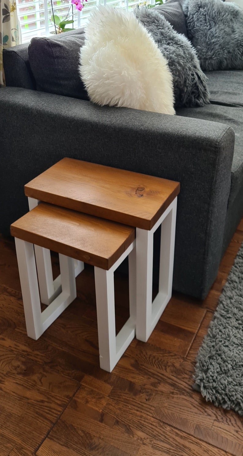 May include: Two wooden nesting tables. The tabletops are a warm brown, and the legs are white. The tables are rectangular and have a simple, modern design. The larger table is approximately 18 inches wide and 18 inches deep.