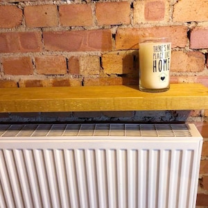 May include: A white candle with the text "There's no place like home" on a wooden shelf against a brick wall. The shelf is above a white radiator.