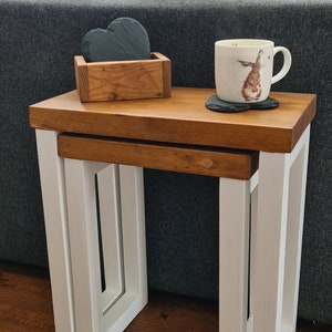 May include: A set of two nesting tables with a wooden top and white legs. The tables are different sizes and are placed one inside the other. A wooden box with heart-shaped coasters and a mug with a rabbit design are on top.