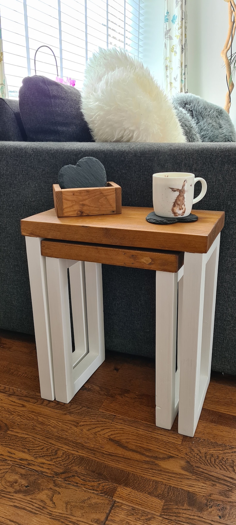 May include: A set of two nesting tables with a wooden top and white legs. The tables are different sizes and are placed one inside the other. A wooden box with heart-shaped coasters and a mug with a rabbit design are on top.