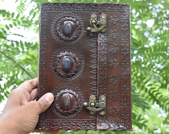 May include: A brown leather journal with a decorative floral design and three blue gemstone accents. The journal has a brass clasp closure.