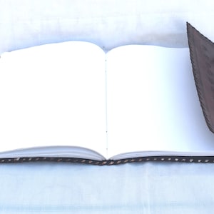 K&ouml;nnte beinhalten: Ein braunes, ledergebundenes Tagebuch mit dekorativen N&auml;hten und Metallschlie&szlig;en. Das Tagebuch ist ge&ouml;ffnet und zeigt leere wei&szlig;e Seiten.