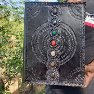 K&ouml;nnte beinhalten: Schwarzes Lederjournal mit einem detaillierten gepr&auml;gten Design mit einem Kreis-Muster und sieben bunten Edelsteinen.
