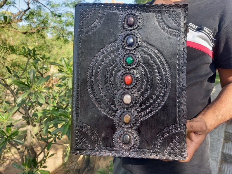K&ouml;nnte beinhalten: Schwarzes Lederjournal mit einem detaillierten gepr&auml;gten Design mit einem Kreis-Muster und sieben bunten Edelsteinen.