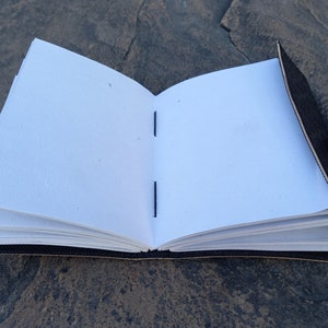 Op de afbeelding: Een zwart lederen gebonden dagboek met een messing sluiting, open om blanco witte pagina's te onthullen die met blauwe draad aan elkaar zijn genaaid.